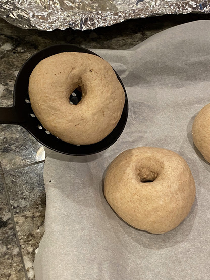 Bagel ready to boil in a black plastic flat spoon-like utensil, about to be put in the water. In the background another bagel sits on a parchment paper tray. Bagel ready to boil in a black plastic flat spoon-like utensil, about to be put in the water. In the background another bagel sits on a parchment paper tray.