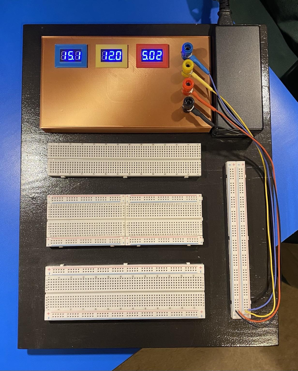a large electronics prototyping board with built in 15, 12, and 5 volt power sources in a 3d-printed gold case on top, colour coded with digital displays showing the voltages provided. Below that are three plugboards (two large, one medium) and a long mini-board serving as power rails going down the right side. It’s all backed by a shiny black backboard in metallic dark grey, several coats of hammered texture paint on quarter-inch plywood. The power cord goes out the top side in back.