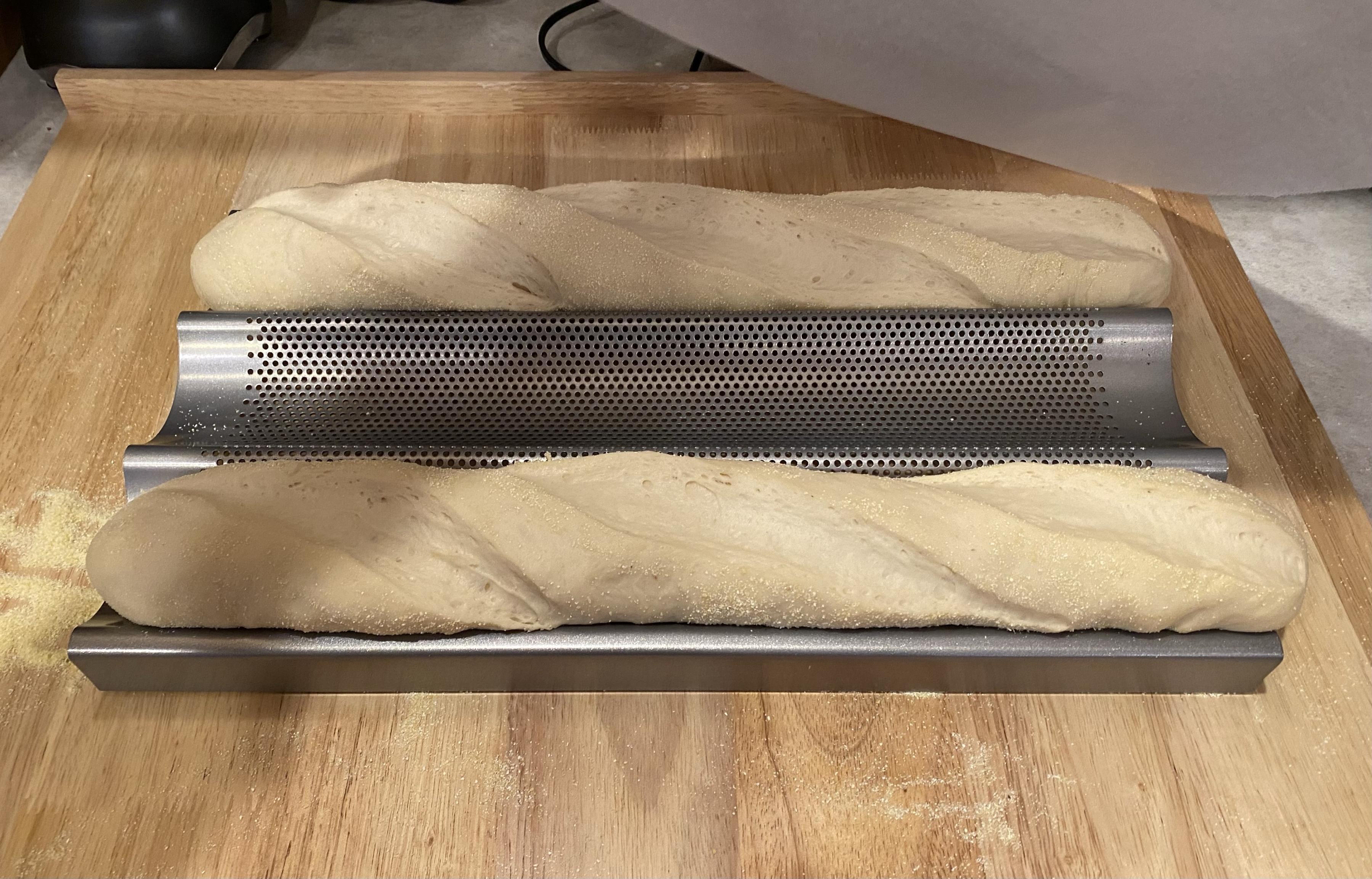 two risen raw baguettes ready to go intk the oven sitting in their baking rack atop a kneading board with some corn meal dust scattered about too