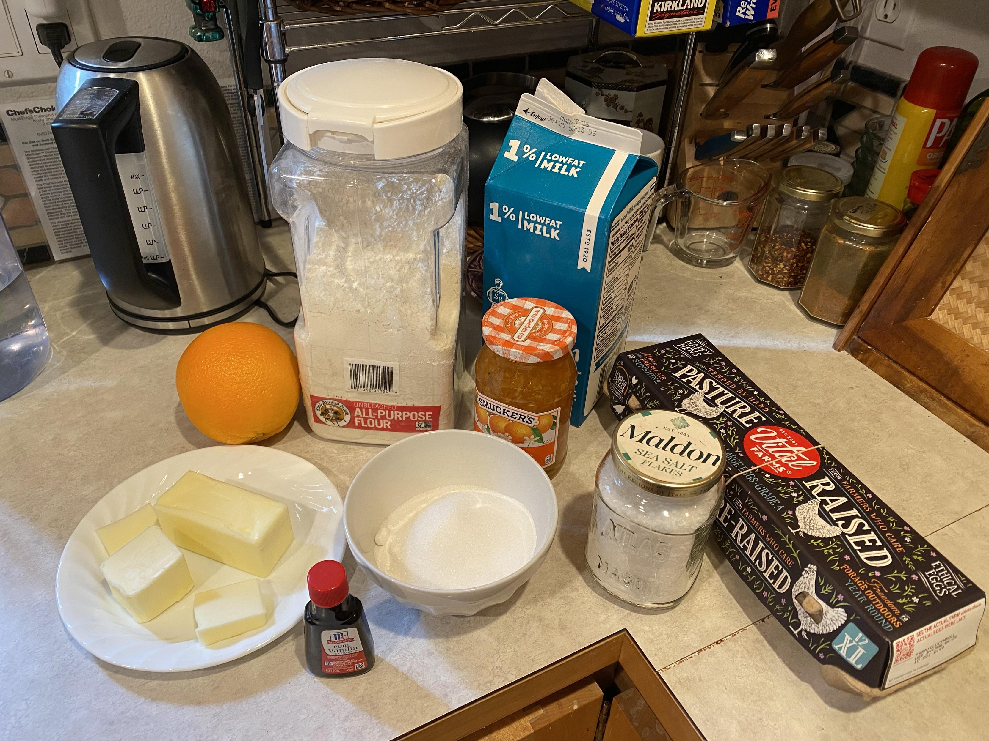 Ingredients for Gateau Breton / Breton Butter Cake on a countertop: flour, milk, orange, marmalade, eggs, flake sea salt, sugar, butter, vanilla extract