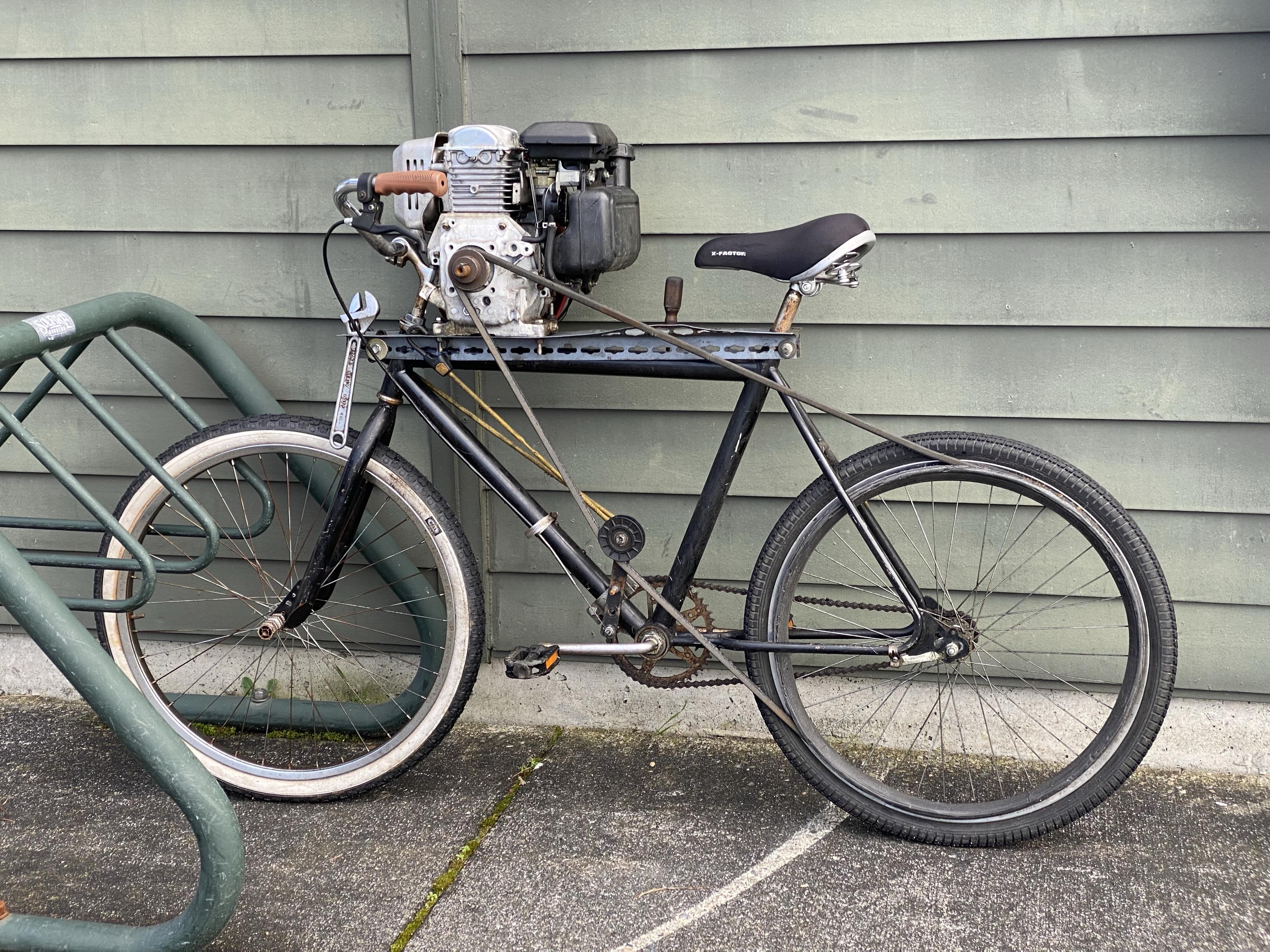 a bicycle in a bike rack at lake forest park city hall with a small two-cycle engine right up against the handlebars and a big belt going from the drive shaft to the rear wheel, one hand brake being a throttle and the other being maybe a drag brake as it affects tension on the belt.
