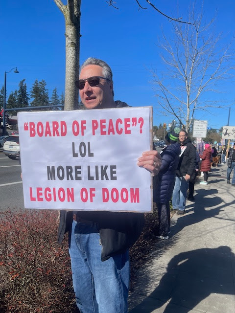 An older white man in sunglasses standing next to a tree on a sidewalk as part of a protest holding up a sign reading "BOARD OF PEACE"? MORE LIKE LEGION OF DOOM on a clear sunny day in Shoreline, Washington.