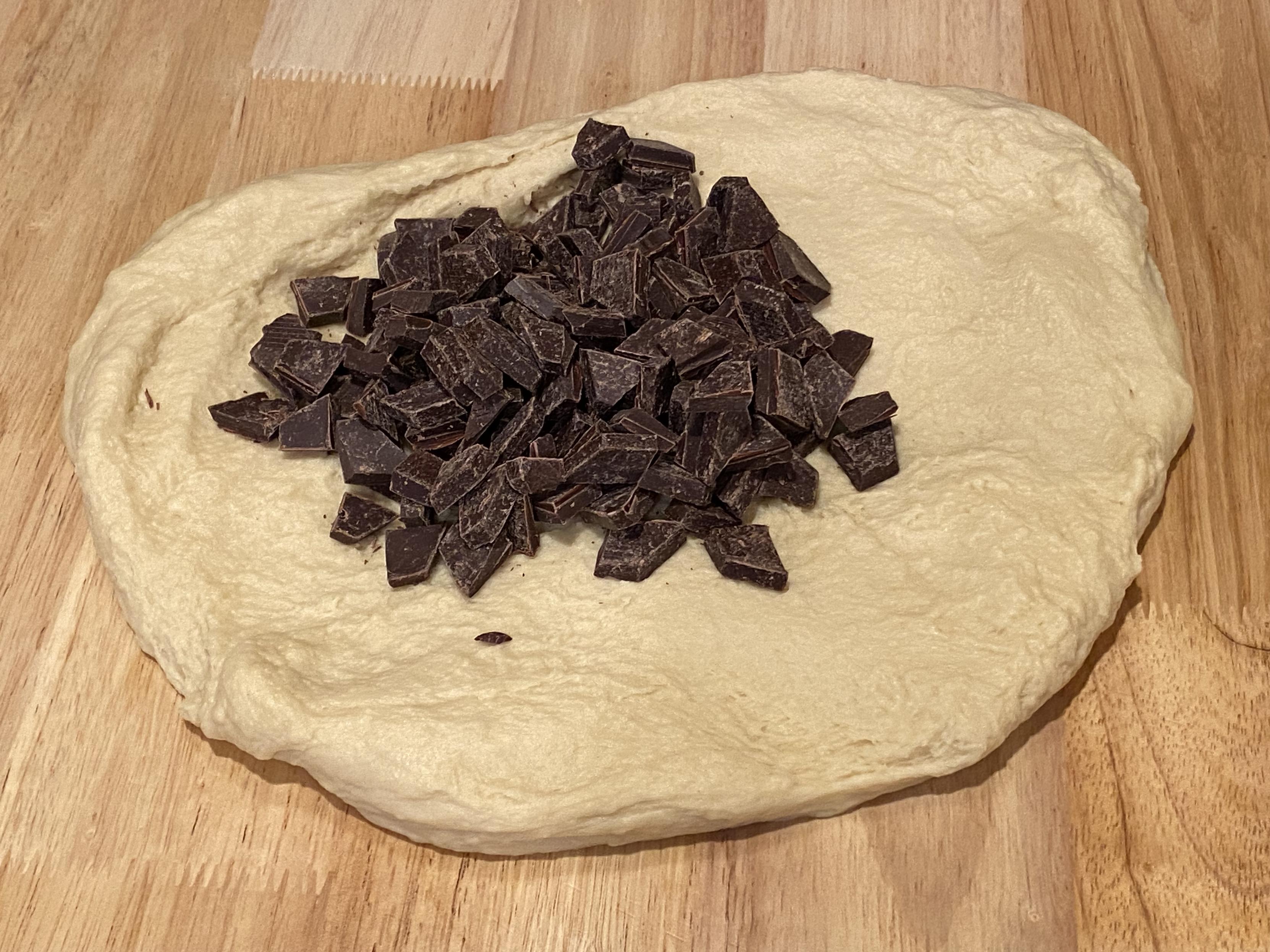 The dough, kneaded out on a board, spread with chocolate chunks dumped atop it