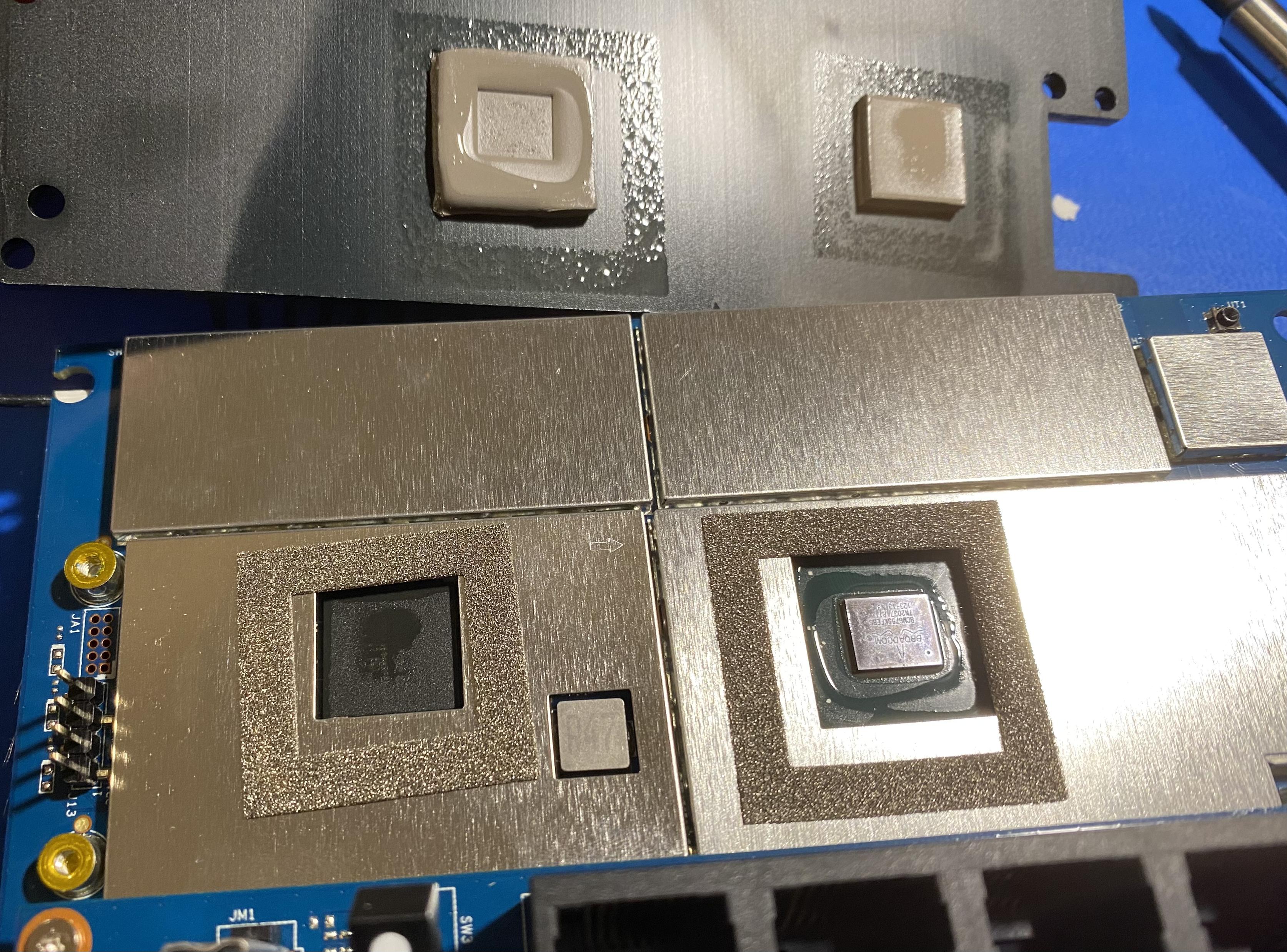 One side of an ASUS router's mainboard with heat sink removed. Gaps in metal coverings where what are presumably processors are exposed have heat pads on the heat sink. Both heat pads, both chips, and the foam areas around said chips are all wet with some sort of fluid.