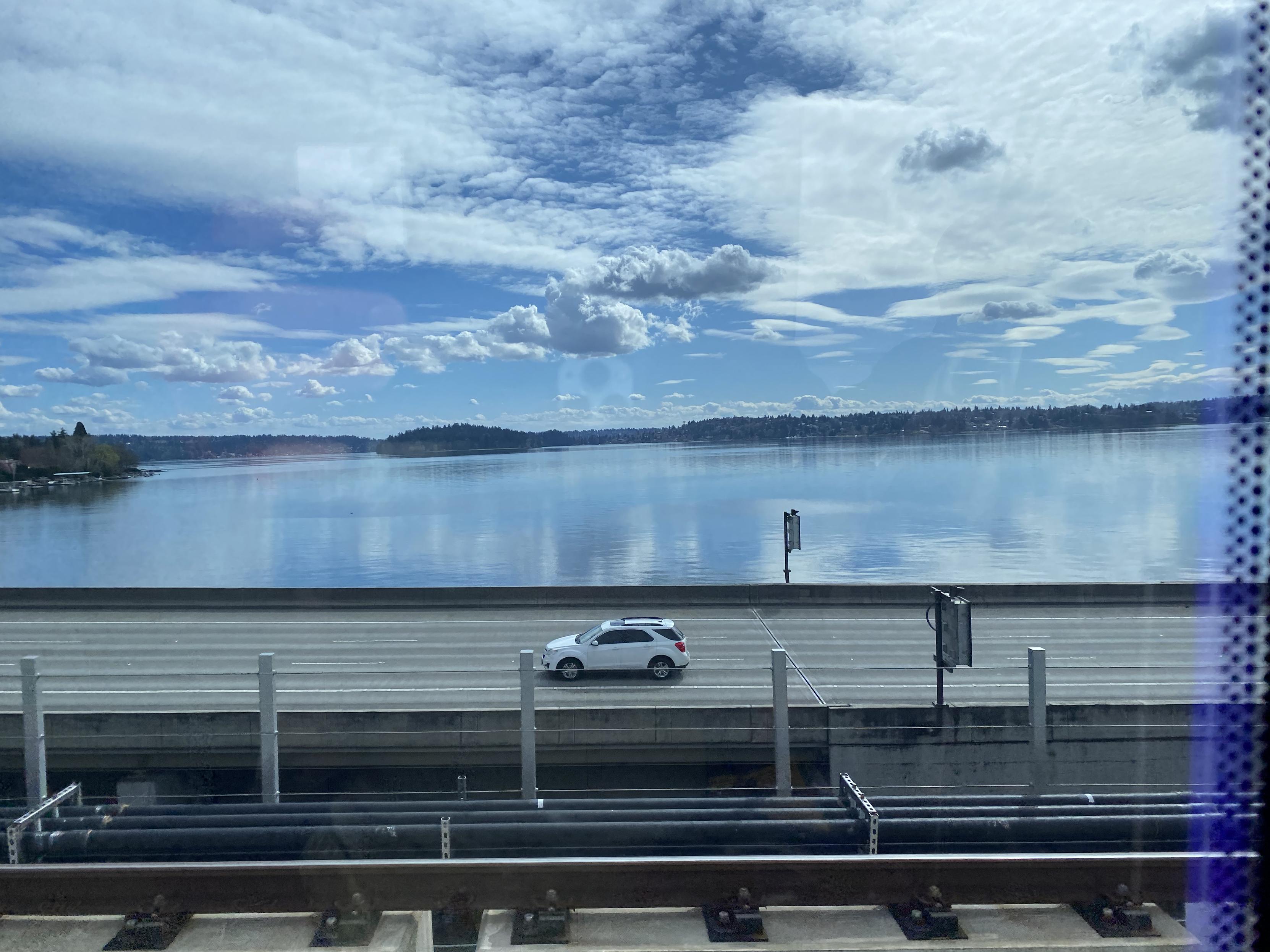 A shot on a partly-cloudy day from a moving light rail car on the Lake Washington Floating Bridge connecting Mercer Island to Seattle of the car lanes on the other set of pontoons and the lake beyond. Past that, you can see down to Renton, Washington.