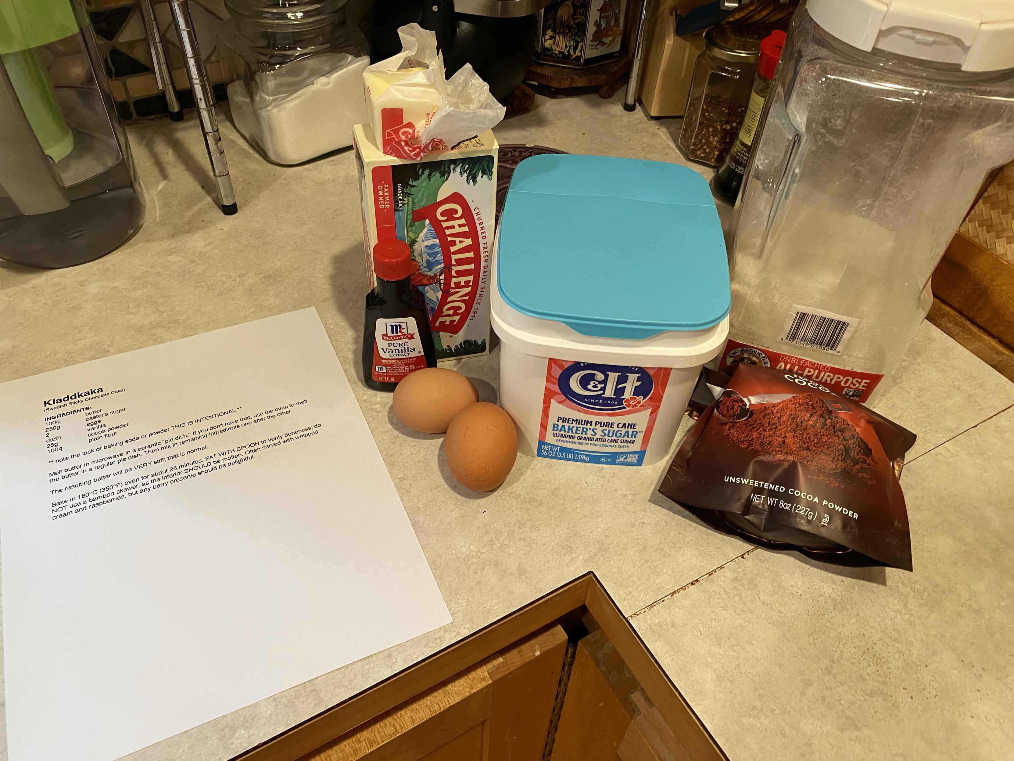 a printed recipe, butter, vanilla extract, eggs, caster’s sugar/baker’s sugar (same thing, still granulated but smaller grains), flour, and cocoa powder all on a countertop