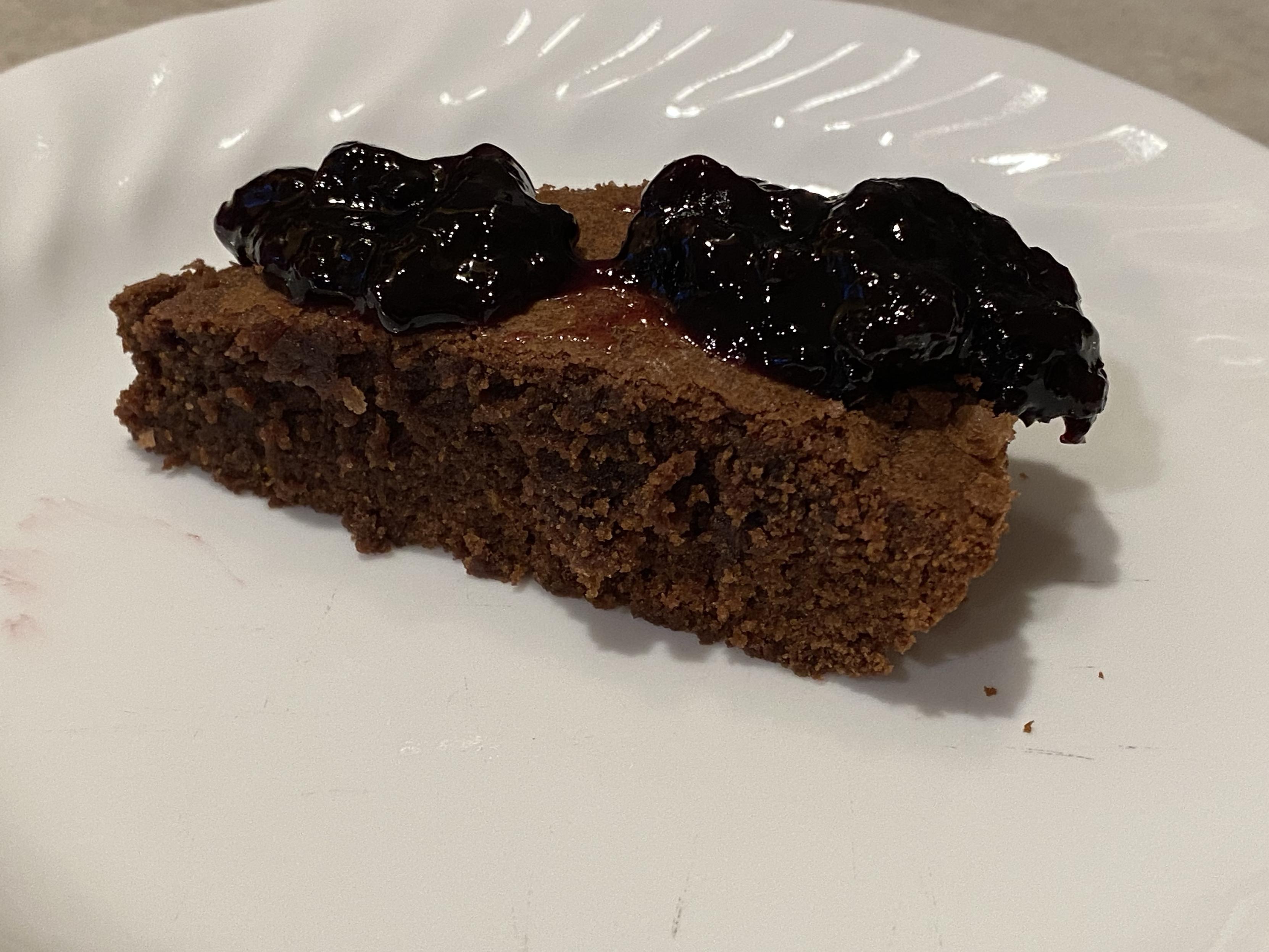 A slice of choco kladdkaka, a brownie-like Swedish cake, topped with blackberry preserves on a white plate. It has a definite dense grain much denser than cake, and a good moistness.