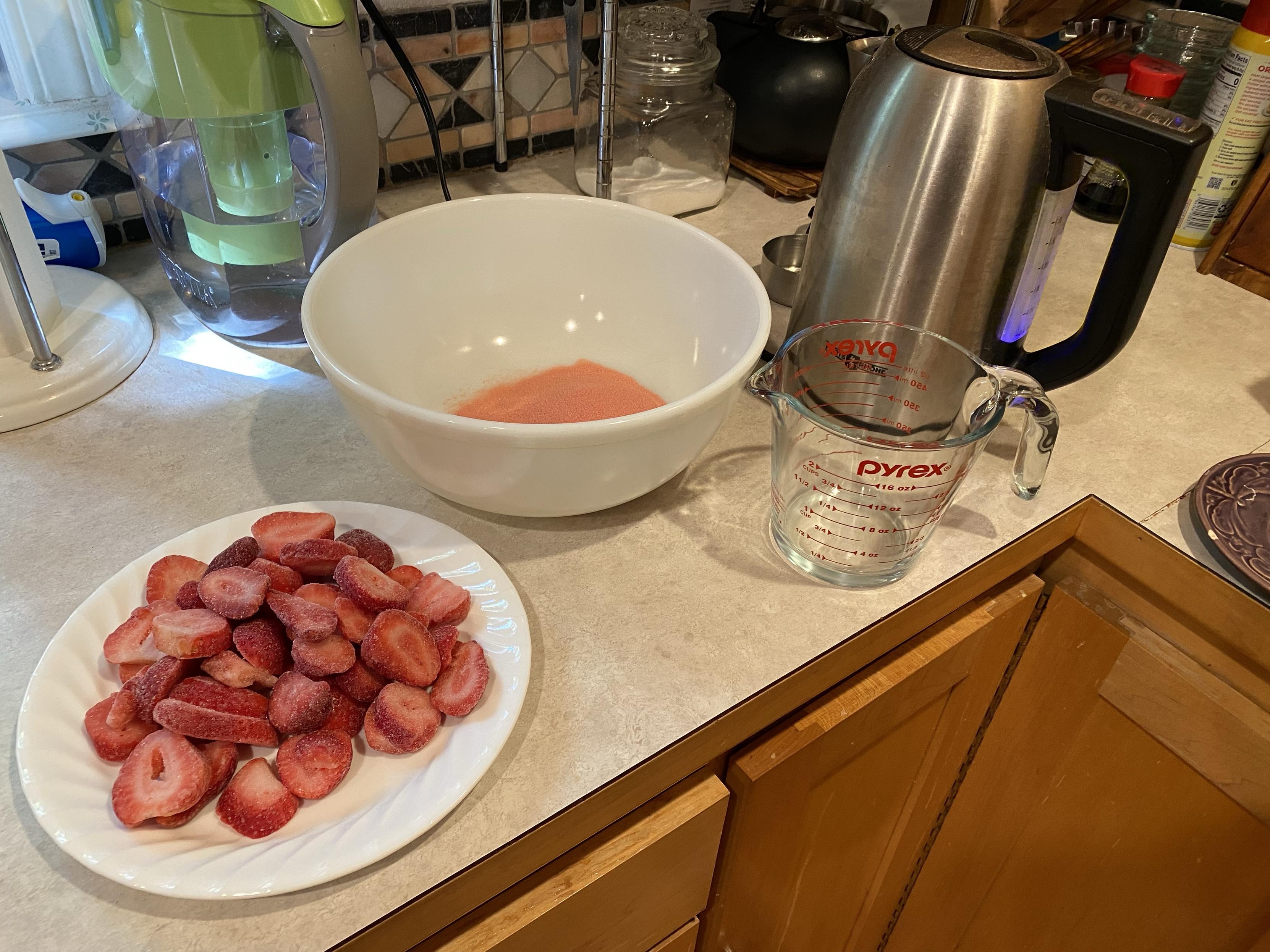 a plate of frozen strawberry slices and a bowl of strawberry jello powder and a kettle and a pyrex measuring cup on a counter. the kettle is ON