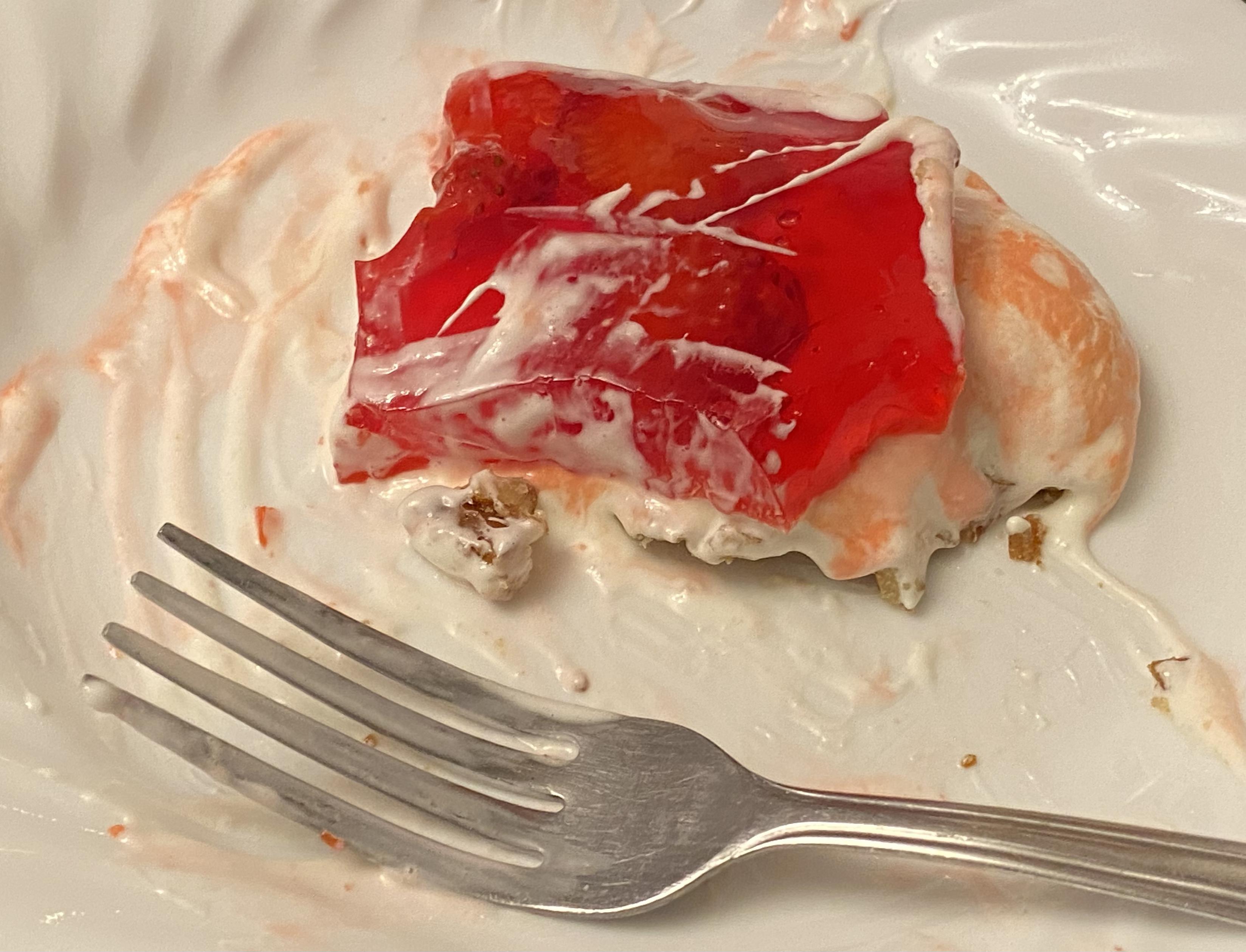 A slice of the pretzel-cream-strawberries-and-jello "salad" dessert , with pieces removed (and eaten), on a white plate with a fork.
