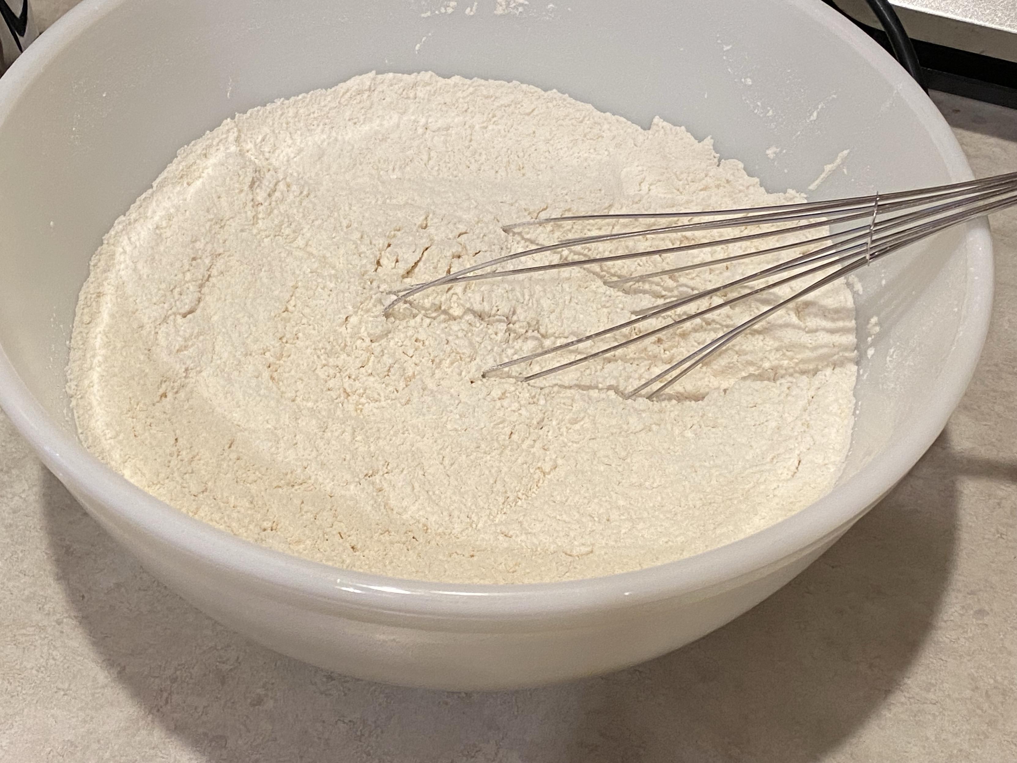 a big whilte bowl filled with flour salt baking soda and powder but mostly flour  and also a whisk