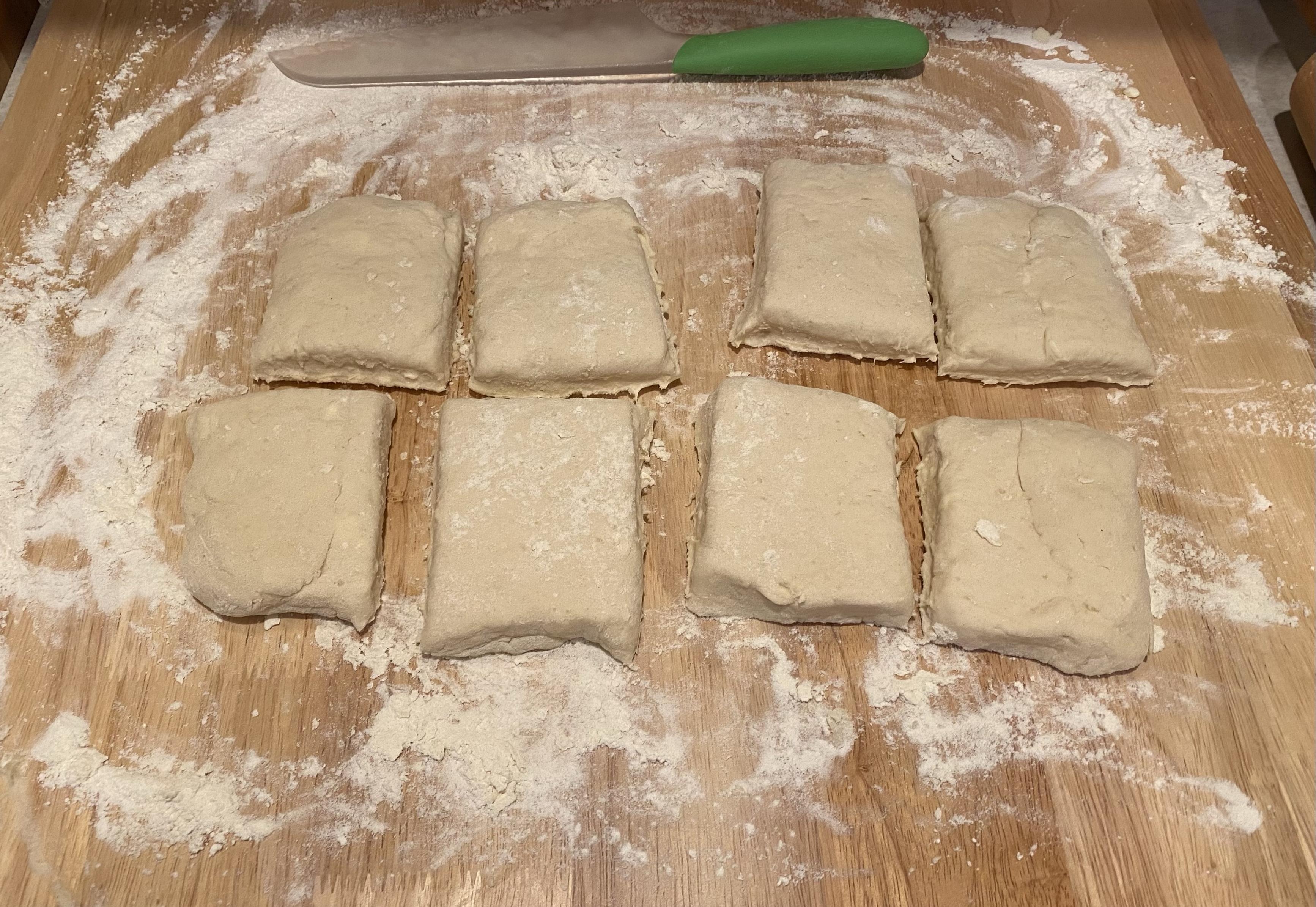eight fairly rectangular biscuit dough cuts on a kneeding board surrounded by board flour and a plastic dividing knife
