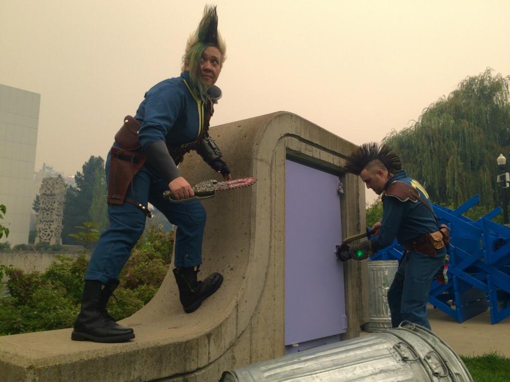 Under a pale yellow sky of smoke, two Wastelanders from Fallout 3 use their weapons to try to break into some sort of vault entrance on the surface. It's a small entry, so is it an escape hatch? Storage? No one knows.