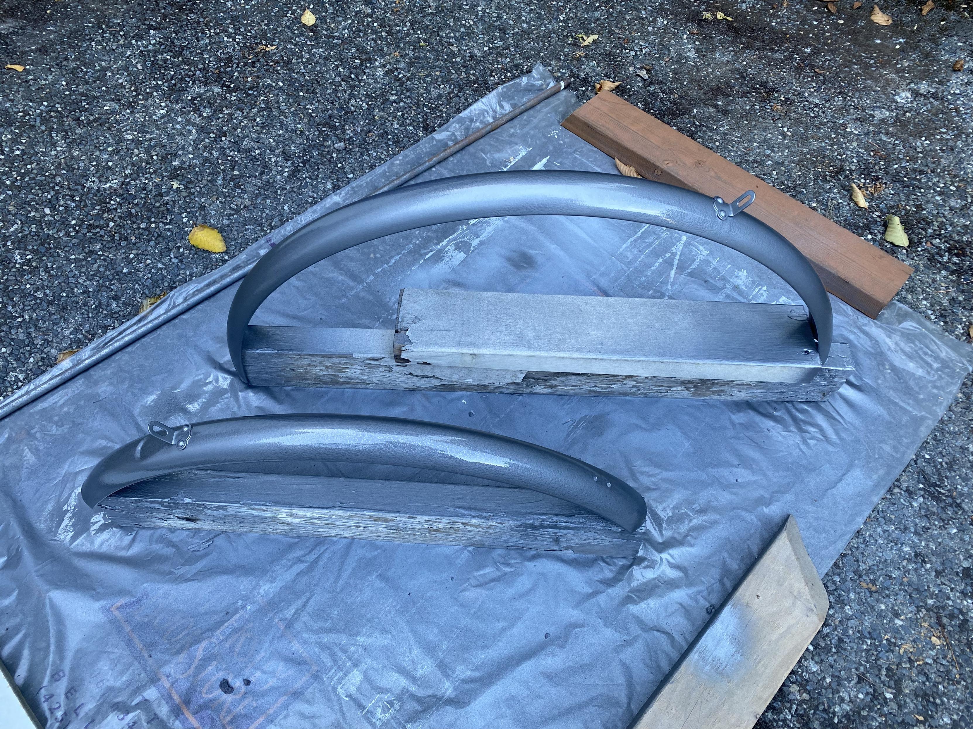 Three-quarter view of the two fenders having just received their fifth and final coat of hammer-texture anti-rust paint in metallic grey, sitting atop a university bookstore bag doubling as a drop cloth, held in place by 2x4s.